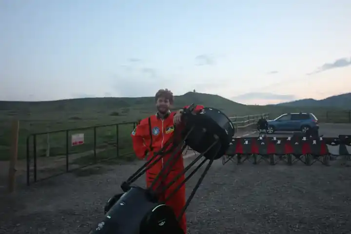 An AstroTours.org astronomer standing beside a large Dobsonian telescope at an outdoor observing site in an illustrative field photograph.